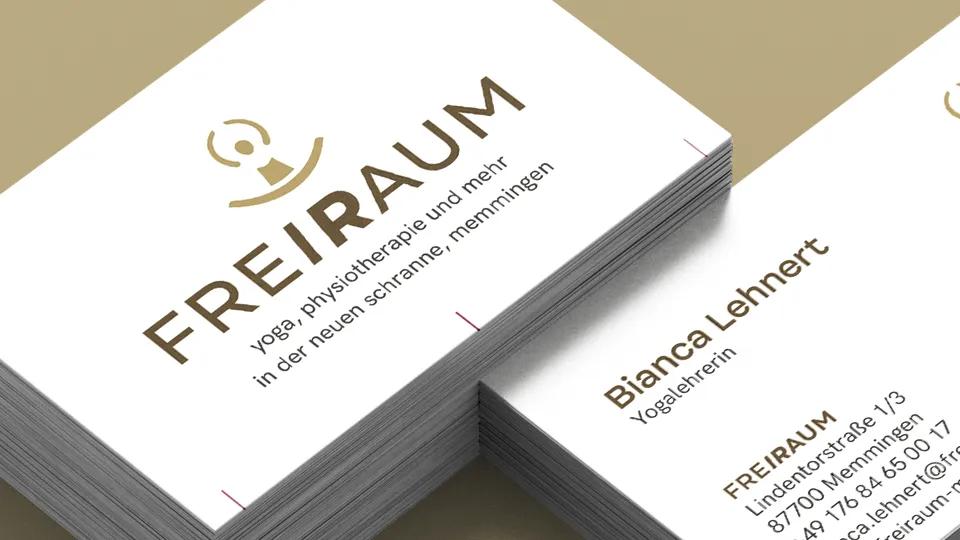 Nahaufnahme eines Stapels hochwertig gedruckter Visitenkarten für 'FREIRAUM', ein Yoga- und Physiotherapie-Studio in Memmingen – gestaltet von einer Corporate Design Agentur für eine einheitliche Markenidentität. Das elegante Design in Braun-Gold-Tönen vermittelt Ruhe und Exklusivität. Das Logo zeigt eine stilisierte, meditierende Figur mit kreisförmigen Linien darüber. Der Name 'FREIRAUM' ist in Großbuchstaben gesetzt, wobei 'RAUM' durch eine abgewandelte Schriftstärke betont wird. Darunter steht die Beschreibung 'yoga, physiotherapie und mehr in der neuen schranne, memmingen'. Diese Visitenkarten sind ein Beispiel für professionelles Corporate Design, das Markenwerte visuell transportiert.