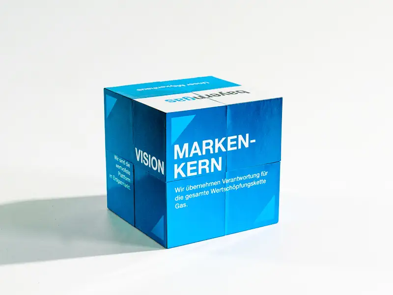 Blauer, faltbarer Würfel mit weißer und hellblauer Schrift, gestaltet im Rahmen eines Corporate Designs durch eine Corporate Design Agentur zur visuellen Darstellung von Unternehmenswerten und Visionen. Auf der sichtbaren Seite stehen die Begriffe 'MARKEN-KERN' und 'VISION' sowie der Text 'Wir übernehmen Verantwortung für die gesamte Wertschöpfungskette Gas.'

Der Würfel hat eine modulare, interaktive Struktur und besteht aus mehreren kleineren Würfeln, die sich neu anordnen lassen. Dieses innovative Design zeigt, wie eine durchdachte Corporate Design Strategie die Markenidentität stärkt und Unternehmenswerte visuell erlebbar macht.
