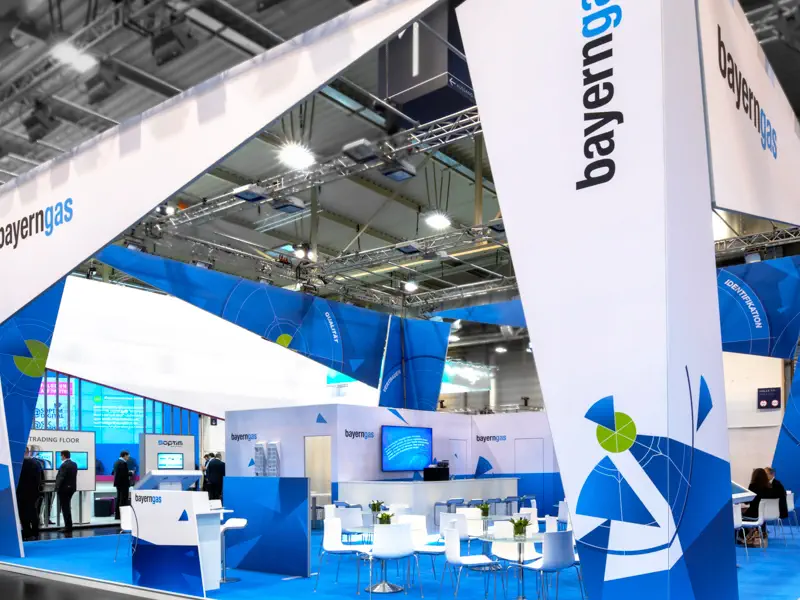 Messestand von Bayerngas mit modernem, offenem Design in Blau und Weiß, entwickelt von einer Corporate Design Agentur zur konsistenten Markenpräsentation. Große, dreieckige Strukturen mit dem Firmenlogo und grafischen Elementen prägen das Erscheinungsbild und schaffen eine starke visuelle Identität.

Im Inneren des Stands befinden sich weiße Tische und Stühle sowie Bildschirme mit Unternehmensinformationen, die das Corporate Design konsequent fortführen. Besucher in dunkler Kleidung sind im Hintergrund zu sehen. Dieses Beispiel zeigt, wie eine durchdachte Corporate Design Strategie für eine einheitliche, professionelle Markenkommunikation sorgt – sowohl digital als auch im Raumdesign.