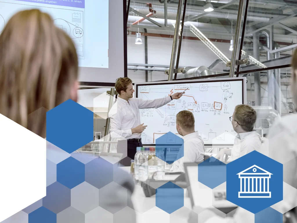 Ein Dozent in weissem Hemd steht vor einem Whiteboard und erklärt ein Diagramm zu Produktionsprozessen. Mehrere Teilnehmer in Business-Kleidung hören aufmerksam zu, einige haben Laptops vor sich. Die Schulung findet in einem modernen, verglasten Schulungsraum mit industrieller Umgebung statt. Überlagert sind blaue hexagonale Formen als Designelemente, rechts unten befindet sich ein blaues Hexagon mit einem weissen Symbol eines Gebäudes, das für Bildung und Schulung steht.
