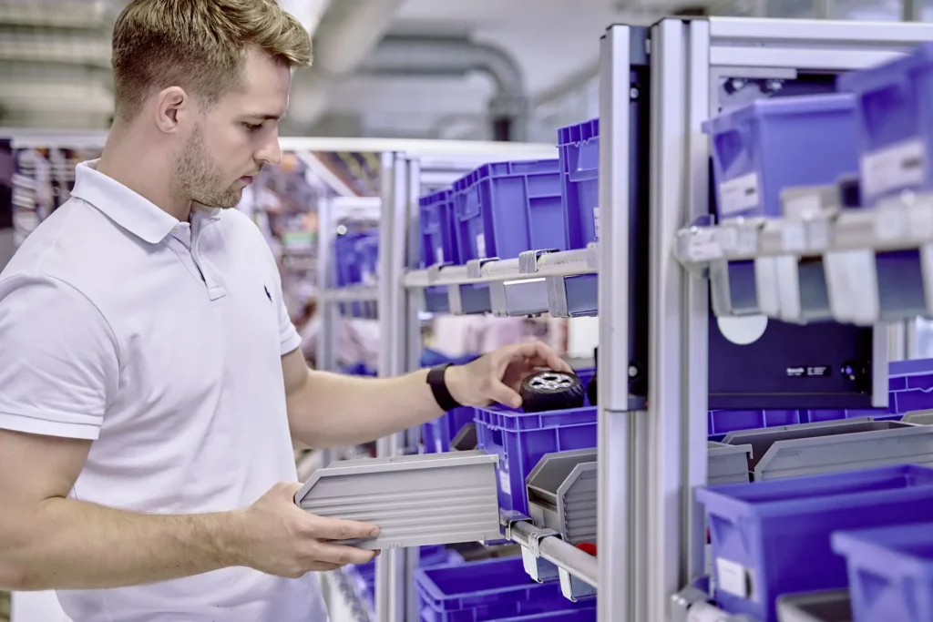 Ein Mann in einem weissen Polohemd steht vor einem Regal mit blauen Kunststoffboxen und entnimmt ein Bauteil, ein Rad mit Felge. In seiner anderen Hand hält er eine graue Ablage. Die Umgebung erinnert an eine moderne Produktions- oder Schulungsumgebung mit einem gut organisierten Materiallager.