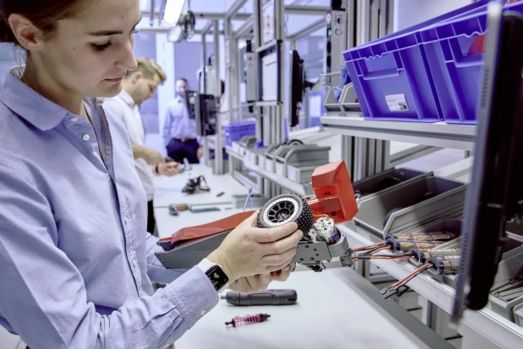 Eine Frau in einem hellblauen Hemd hält ein technisches Modellfahrzeug und setzt ein Rad daran an. Sie trägt eine Smartwatch und arbeitet konzentriert. Im Hintergrund befinden sich weitere Personen an Arbeitsstationen, Regale mit blauen Kunststoffboxen sowie Ablagen mit elektronischen Bauteilen. Die Umgebung erinnert an eine Lernfabrik oder ein Hightech-Labor.