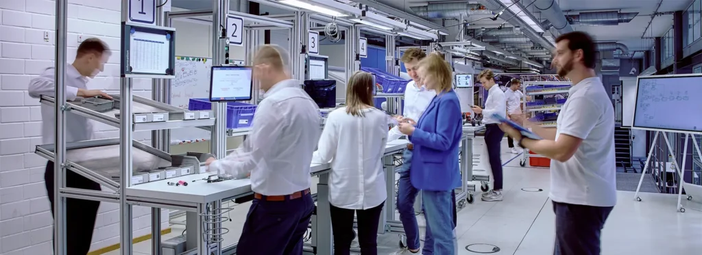 Moderne Lernfabrik mit mehreren Personen, die an Arbeitsstationen in einer Produktionsumgebung tätig sind. Die Halle ist mit nummerierten Stationen, Monitoren und Lagerregalen ausgestattet. Im Vordergrund unterhält sich eine Frau im blauen Blazer mit einem Kollegen, während rechts ein Mann mit Klemmbrett gestikuliert. Im Hintergrund arbeitet eine weitere Person an einem Whiteboard mit Prozessdiagrammen.