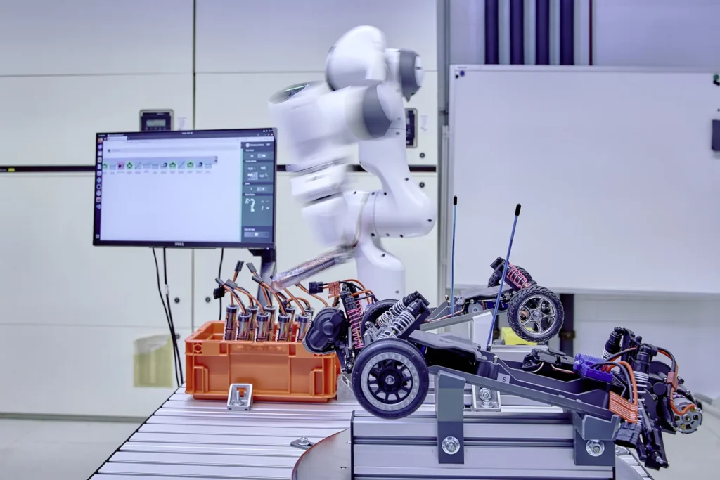 Industrielle Laborumgebung mit einem weissen Roboterarm, der sich in Bewegung befindet und an einer elektronischen Baugruppe arbeitet. Auf dem Tisch stehen ein Modellfahrzeug mit freigelegten Bauteilen sowie eine orangefarbene Kiste mit Kabeln und Komponenten. Im Hintergrund ist ein Monitor mit einer Steuerungssoftware sowie eine weisse Tafel zu sehen.