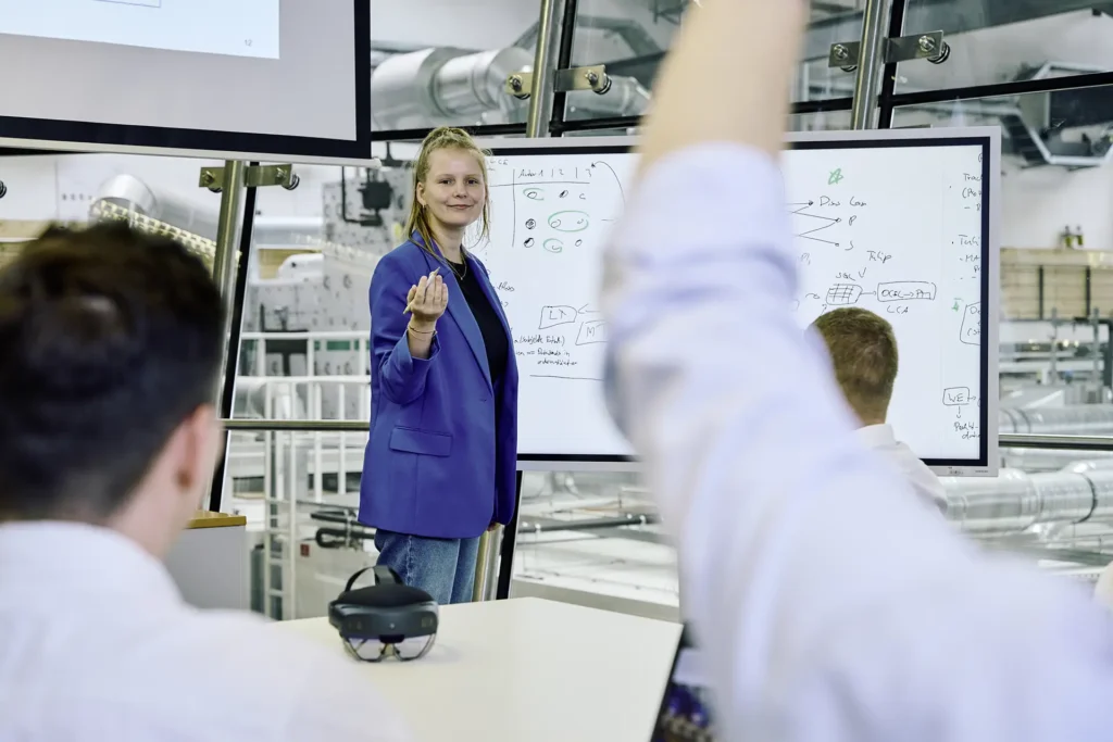 Eine junge Frau in einem blauen Blazer steht vor einem Whiteboard mit handschriftlichen Notizen und Diagrammen und hält eine Schulung oder Präsentation. Sie lächelt und macht eine einladende Geste. Im Vordergrund sitzt ein Teilnehmer mit erhobener Hand, während ein anderer mit dem Rücken zur Kamera zuhört. Auf dem Tisch liegt eine AR-Brille. Der Raum hat eine moderne Glasarchitektur mit Blick auf eine industrielle Umgebung.