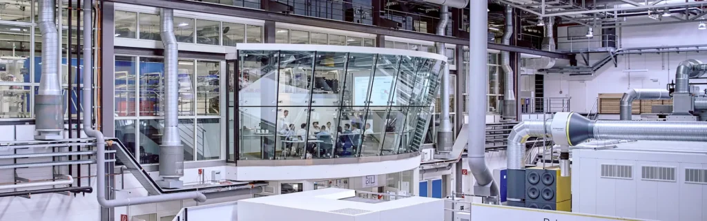 Innenansicht einer modernen Industriehalle mit einer verglasten, schwebenden Besprechungsraum-Struktur. Im Inneren des Raums sitzt eine Gruppe von Personen an einem Tisch, während eine Person vorne steht und eine Präsentation hält. Die Halle ist mit zahlreichen Rohren, Belüftungssystemen und Metallstrukturen ausgestattet, was auf eine hochtechnisierte Fertigungs- oder Forschungsumgebung hindeutet.