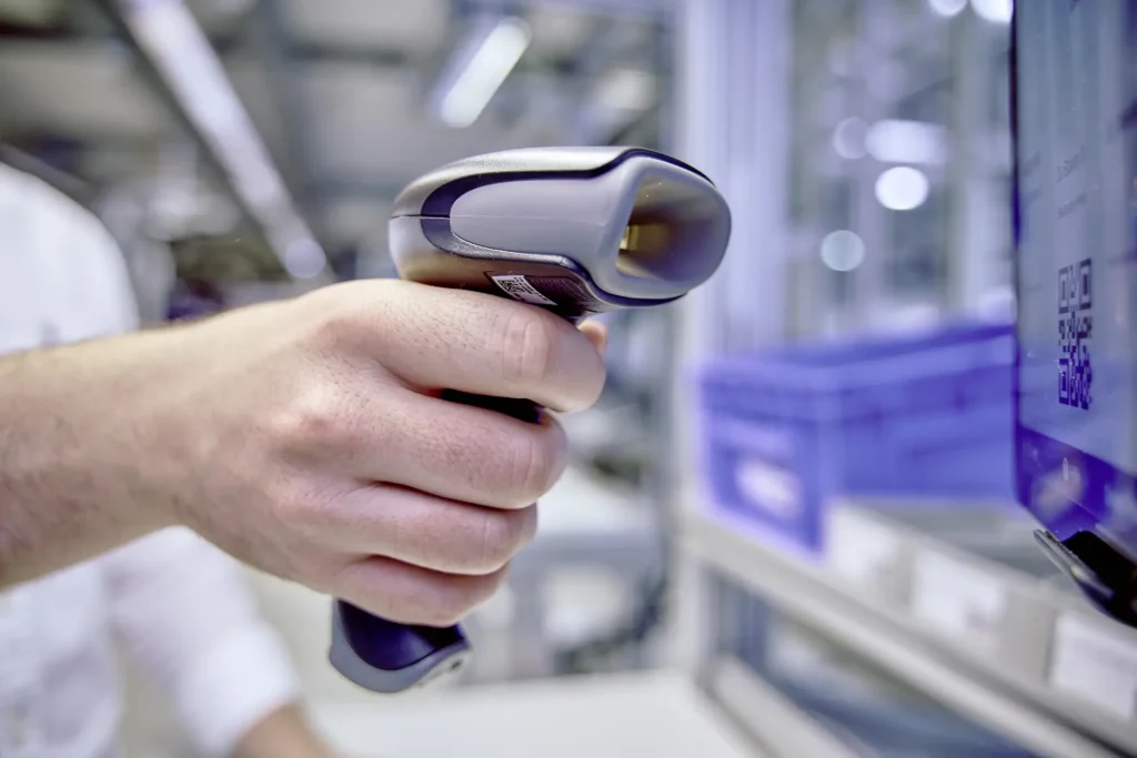 Nahaufnahme einer Hand, die einen Barcode-Scanner hält und auf einen QR-Code auf einem Bildschirm richtet. Der Hintergrund zeigt eine industrielle oder logistische Umgebung mit Regalen und blauen Kunststoffboxen. Die Szene vermittelt eine moderne, digitalisierte Arbeitsumgebung.