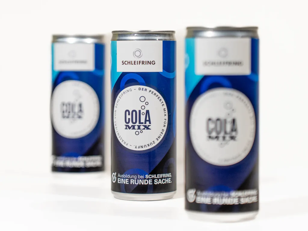 Drei Cola-Mix-Dosen mit individuellem Design für die Firma SCHLEIFRING. Die Dosen sind blau mit einem weissen Kreis in der Mitte, in dem „COLA MIX“ steht, umgeben von der Aufschrift „Der perfekte Mix für deine Zukunft“. Oben befindet sich das SCHLEIFRING-Logo, unten der Slogan „Ausbildung bei SCHLEIFRING. EINE RUNDE SACHE.“ Die vorderste Dose ist scharf fokussiert, die anderen zwei sind leicht verschwommen im Hintergrund.