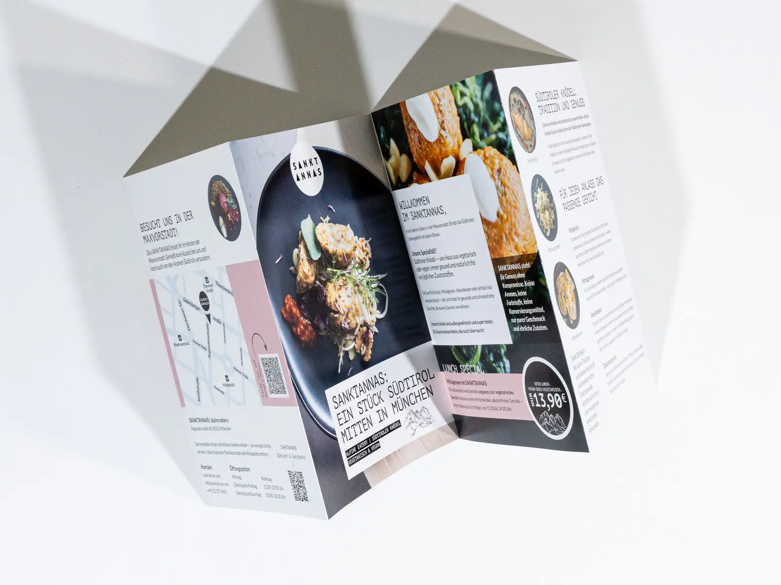 Geöffneter Faltprospekt des Restaurants „Sankt Annas“ in München. Das Design kombiniert Weiss-, Schwarz- und Rosatöne mit Bildern von Speisen, darunter ein Gericht auf einem schwarzen Teller. Die Broschüre enthält Informationen zur Philosophie des Restaurants, zur Spezialität „Südtiroler Knödel“, zur Mittagskarte sowie eine Standortkarte mit Adresse und QR-Code. Ein Preis von „13,90 €“ ist für ein Menü ersichtlich. Die Gestaltung hebt das Thema Alpenküche und moderne Gastronomie hervor.
