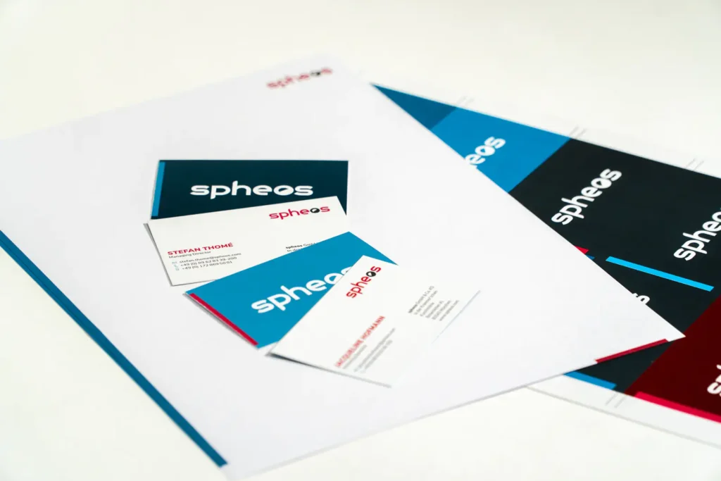 Geschäftsausstattung der Marke „spheos“ mit Briefpapier, Visitenkarten und Branding-Elementen. Die Designs nutzen eine moderne Farbpalette aus Blau, Weiß, Rot und Schwarz mit klarer Typografie. Einige Visitenkarten zeigen Namen und Kontaktdaten, während im Hintergrund weitere Drucksachen mit dem „spheos“-Logo sichtbar sind.  
