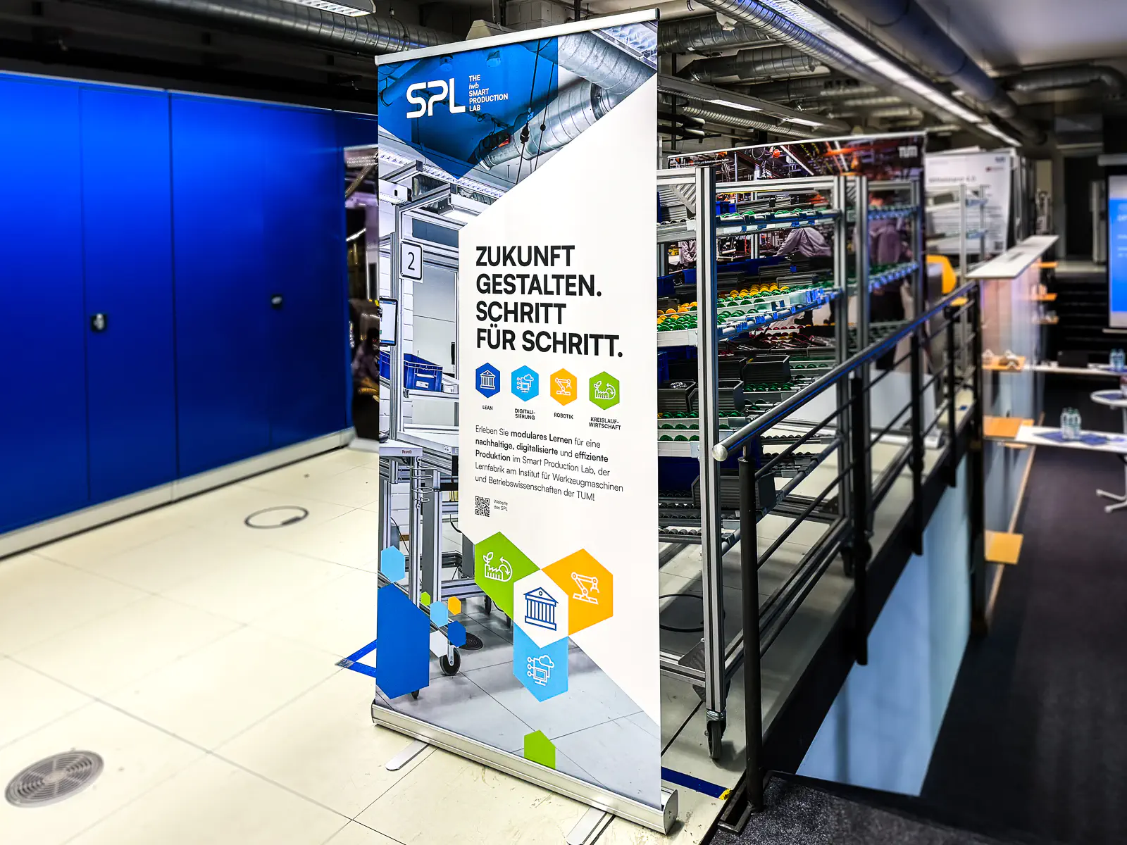 Roll-up-Banner des „Smart Production Lab“ (SPL) in einer industriellen Umgebung. Das Banner zeigt ein modernes Fabriklayout mit Metallregalen und Förderbändern im Hintergrund. Der Text „Zukunft gestalten. Schritt für Schritt.“ hebt die Themen Lean, Control & Automation sowie Kreislaufwirtschaft hervor. Farbige Icons und geometrische Designelemente unterstützen das visuelle Konzept.  

